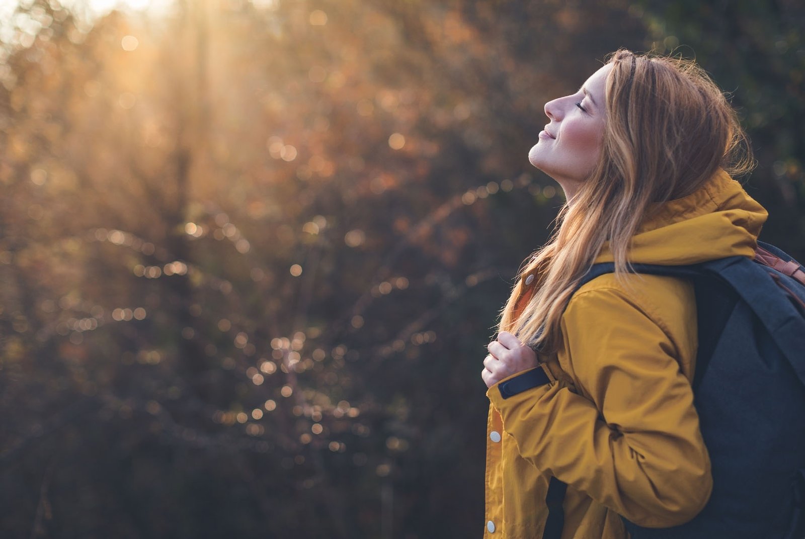 Frau in gelber Jacke geht im Wald spazieren und schaut ins Morgenlicht