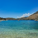 grünblaues Meer mit kleinen Booten in der Ferne vor einer Berglandschaft mit weißen Wölkchen